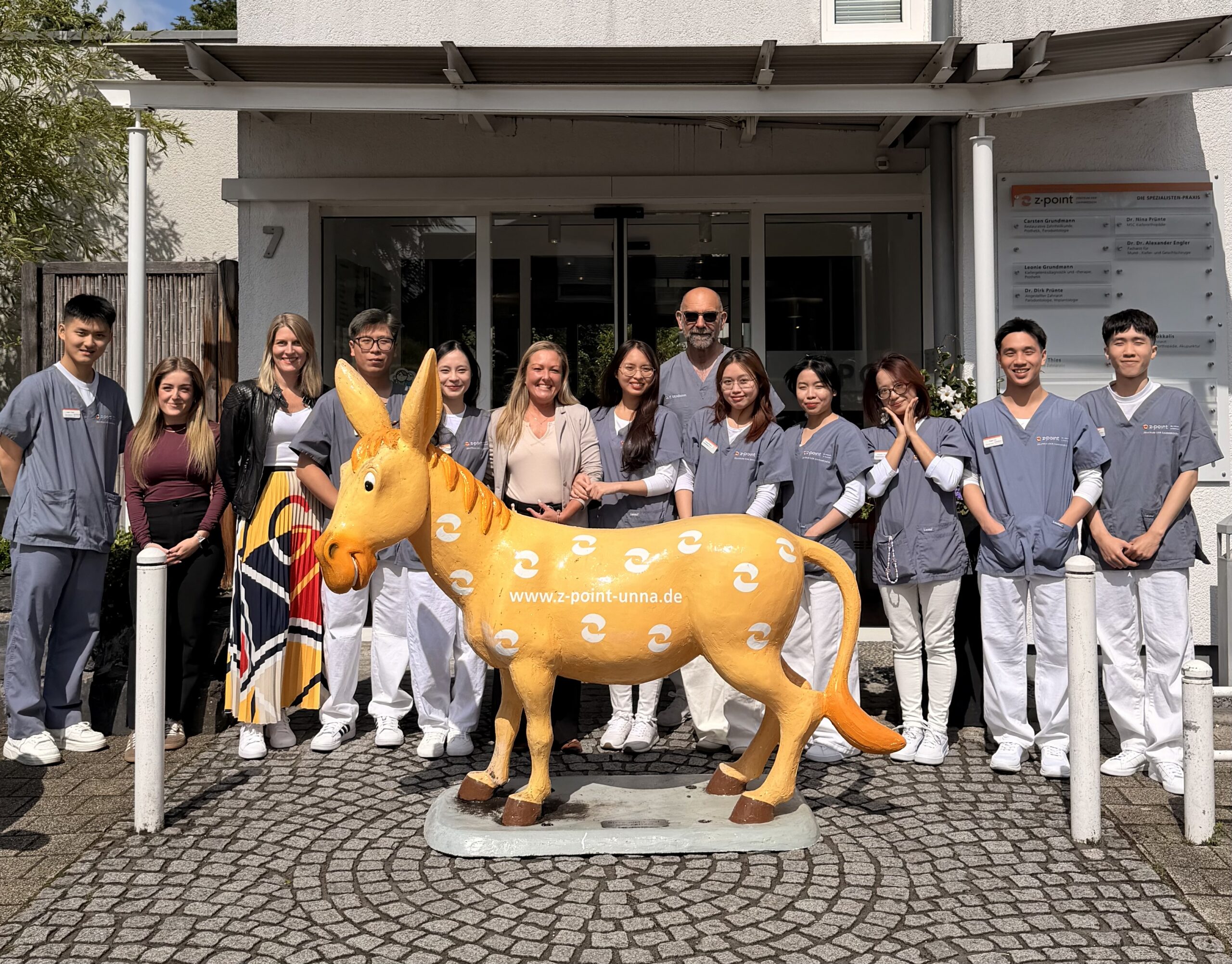 Group of students in scrubs posing with an orange horse statue in front of a building entrance.